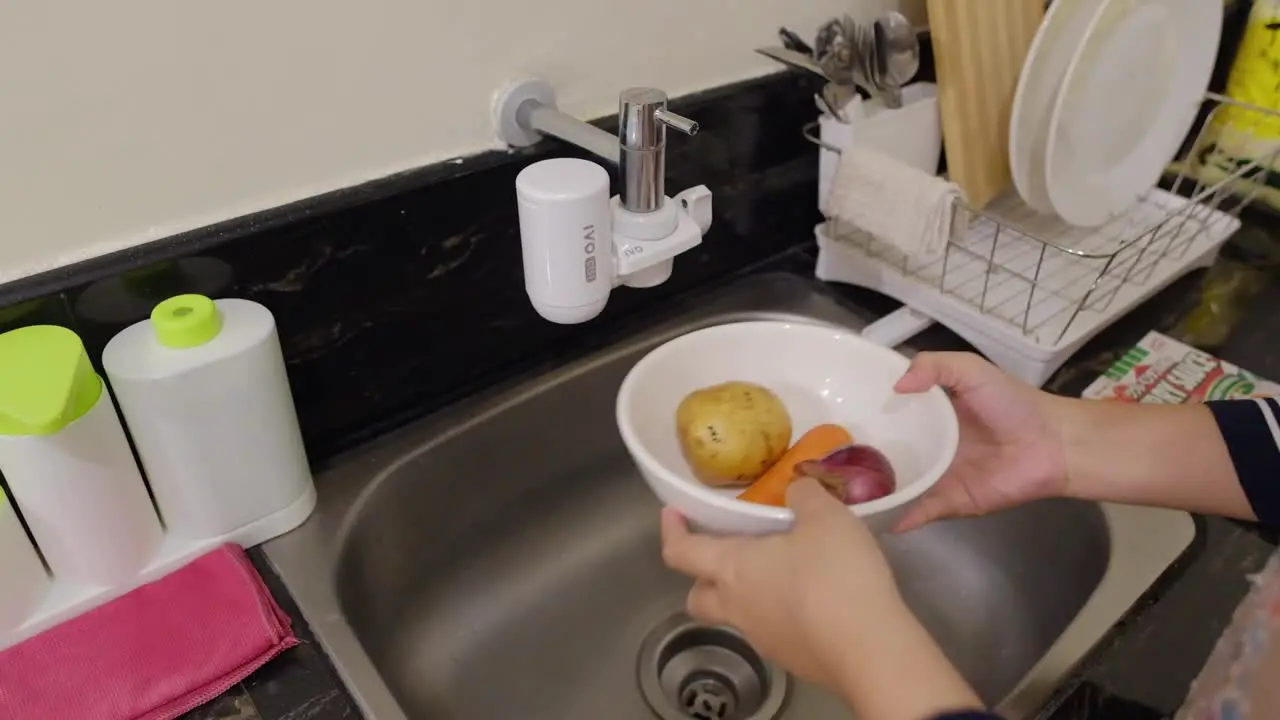 Close-up shot of hands washing vegetables in a bowl under kitchen sink faucet