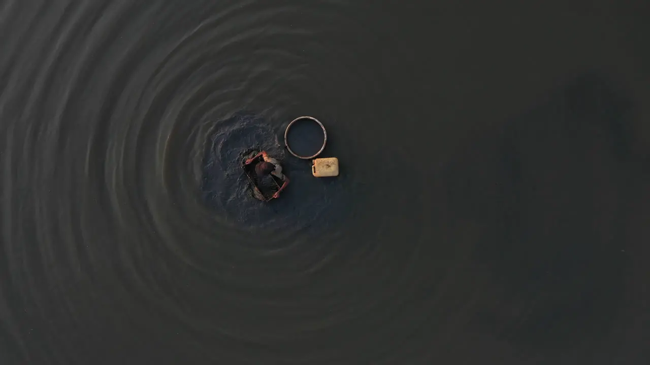 A slow zoom in from above as a man is collecting snails in the muddy shallows of a canal in Vietnam