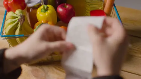 Person Checking Bill Or Receipt For Supermarket Shopping Basket Full Of Basic Food Items