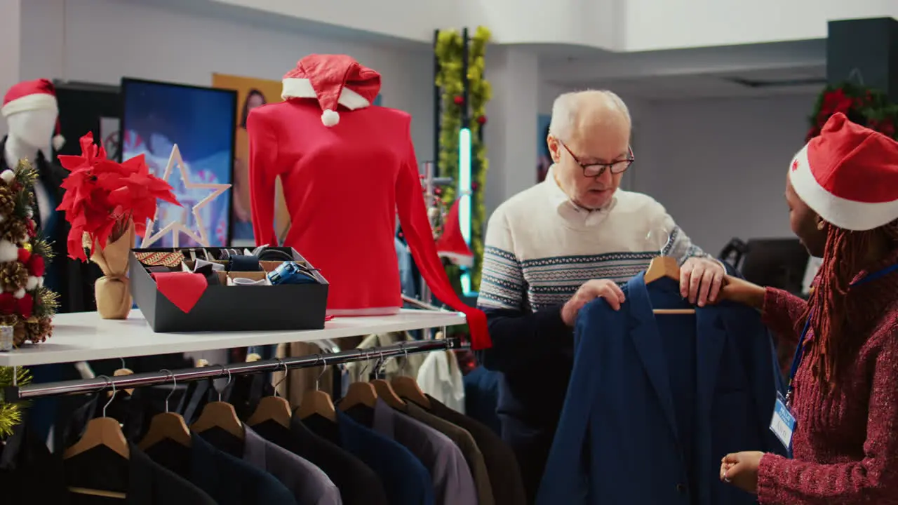 Skillful employee in Christmas decorated clothing shop upselling luxurious blazer to rich senior client convincing him of garment being masterfully crafted with quality materials