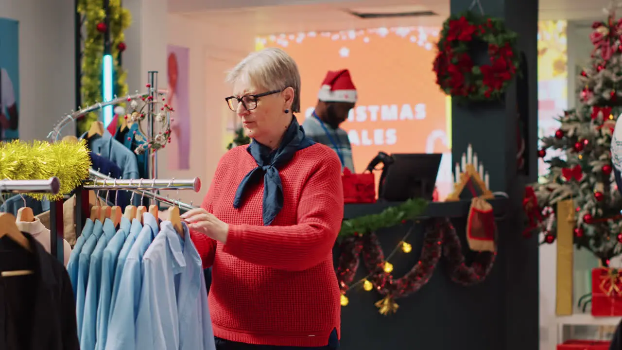 Senior woman browsing through blazer racks in Christmas decorated clothing store while husband pays at shop counter in background Clients making xmas purchases during winter holiday season