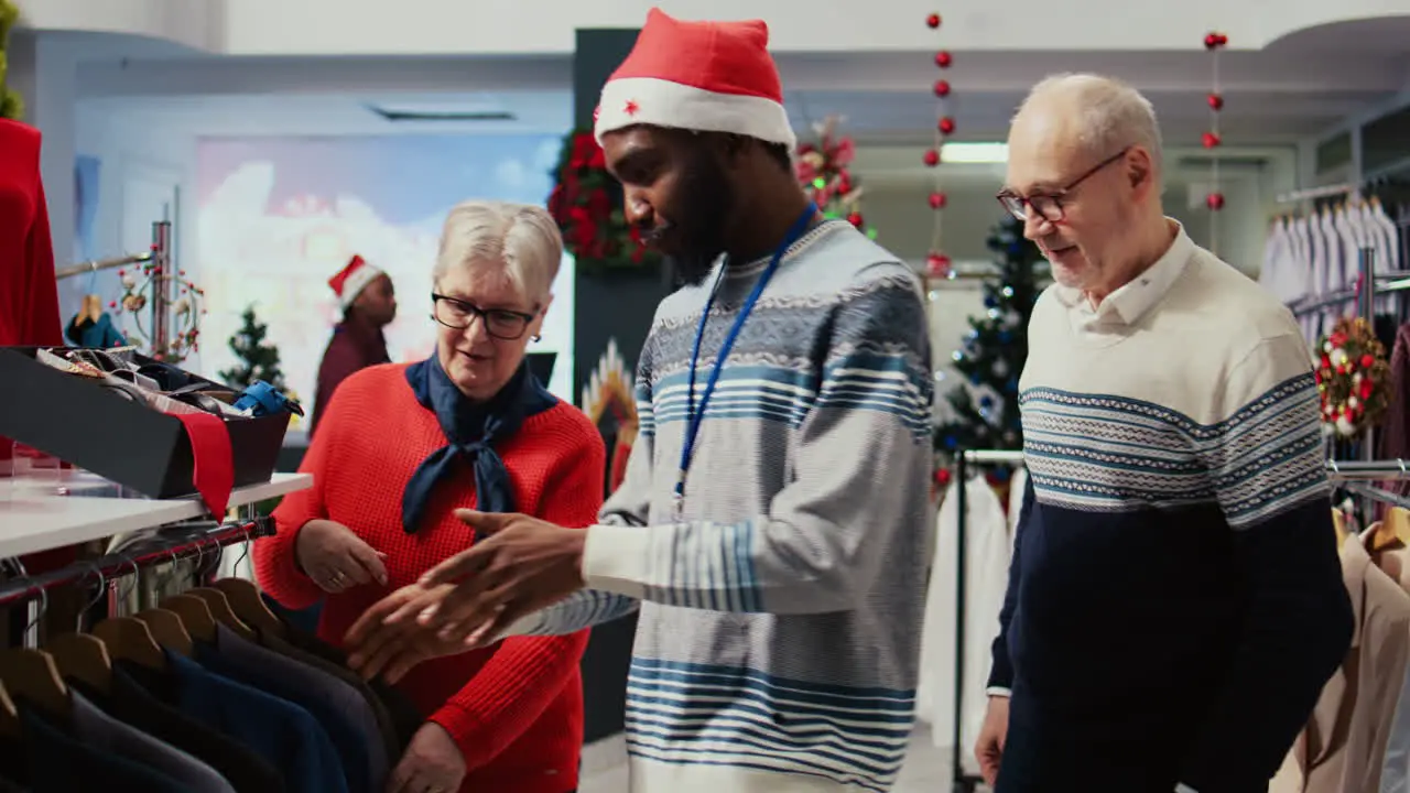 Wife and husband in Christmas decorated shopping mall clothing store being assisted by african american employee with finding perfect outfit during winter holiday season promotional sales