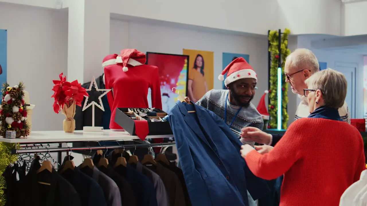 Wife and husband in xmas ornate shopping mall fashion shop being assisted by african american worker with finding ideal outfit during festive holiday season promotional sales