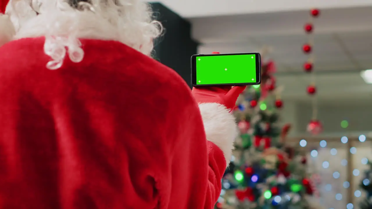 Employee in festive ornate shopping mall wearing Santa Claus suit holding green screen phone taking a break from job shift and watching videos on chroma key screen close up