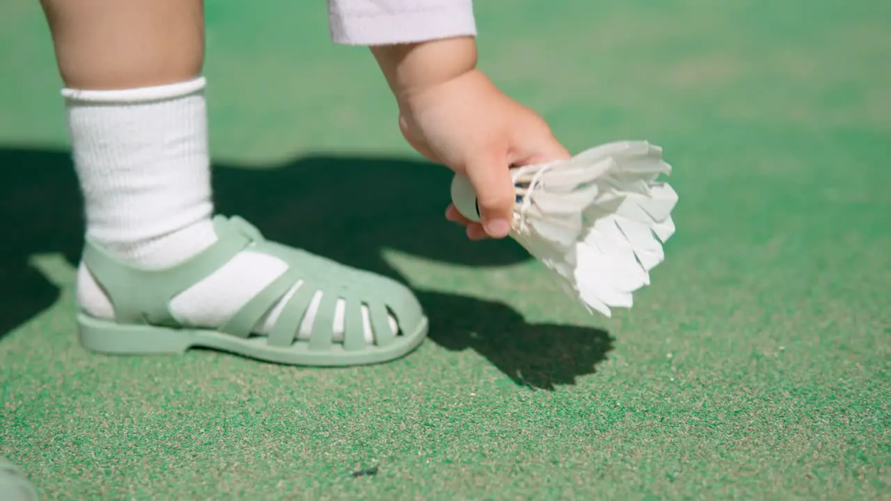 Toddler's hand picking up white badminton shuttlecock from the ground of green court outdoors close-up