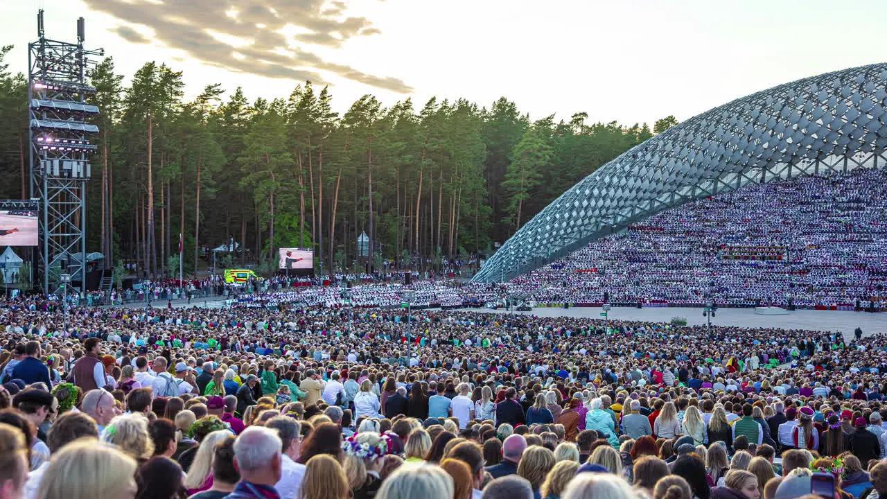 Timelapse shot of huge crowd of locals gathering to watch the Latvian Song and Dance Festival in an outdoor stadium in Riga Latvia with sun setting in the background