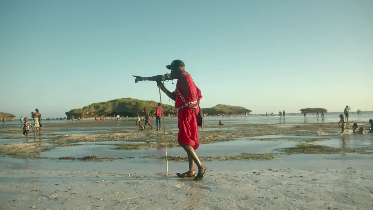 Masai Warrior In Red Traditional Clothes Standing At The Beach Holding A Wooden Pole And Pointing Direction In Watamu Kenya