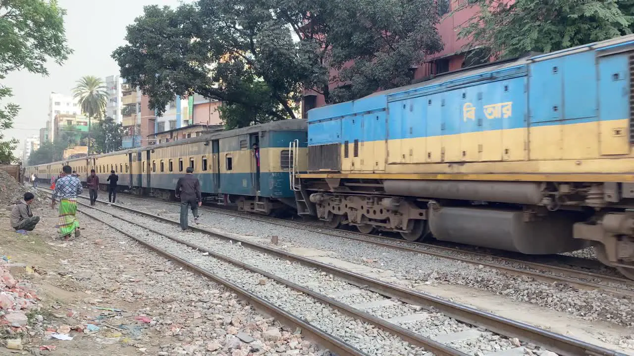 Bengali Male Carrying Basket On Head Walking Past Railway Tracks With Train In Background In Dhaka