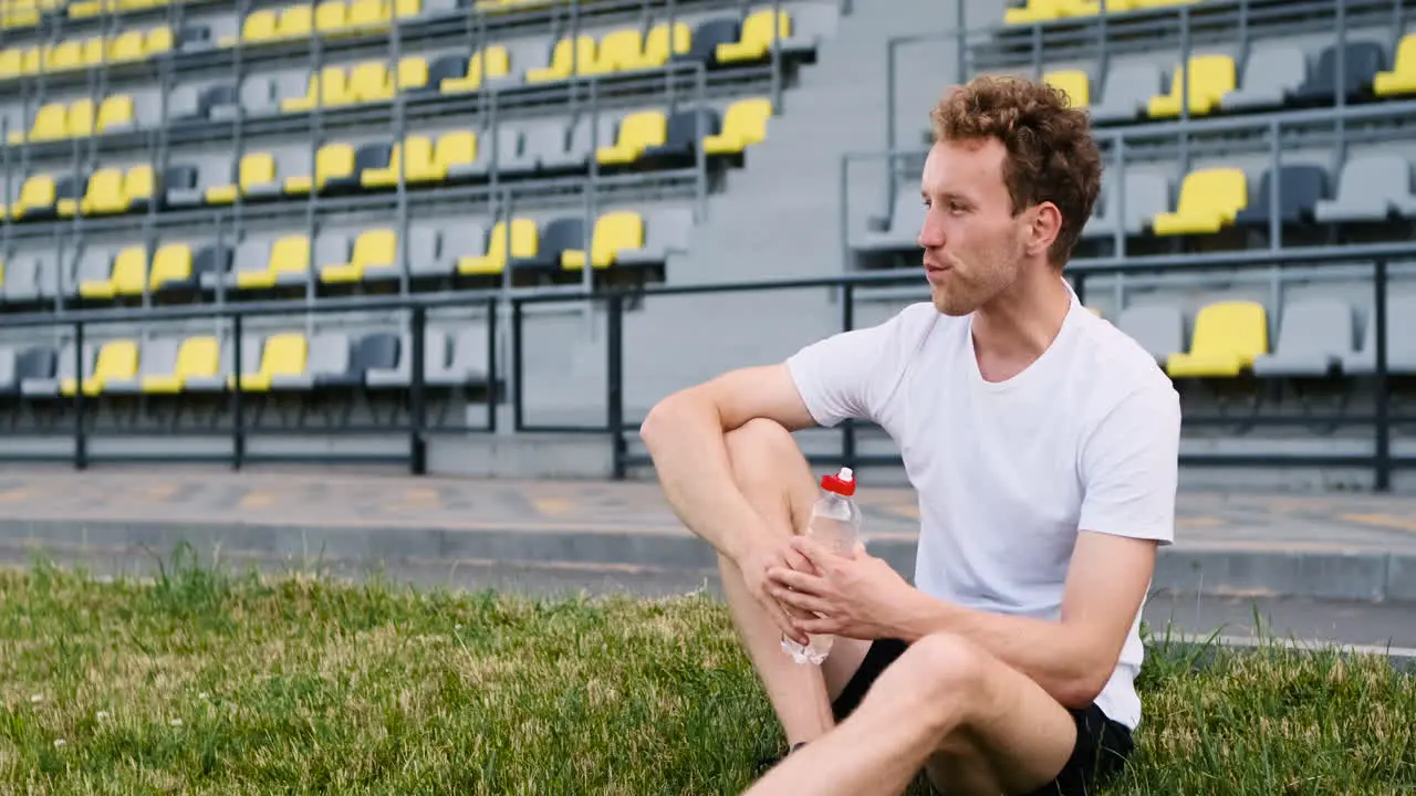 Sportive Man Resting And Drinking Water While Sitting On The Grass In A Stadium After Running Workout