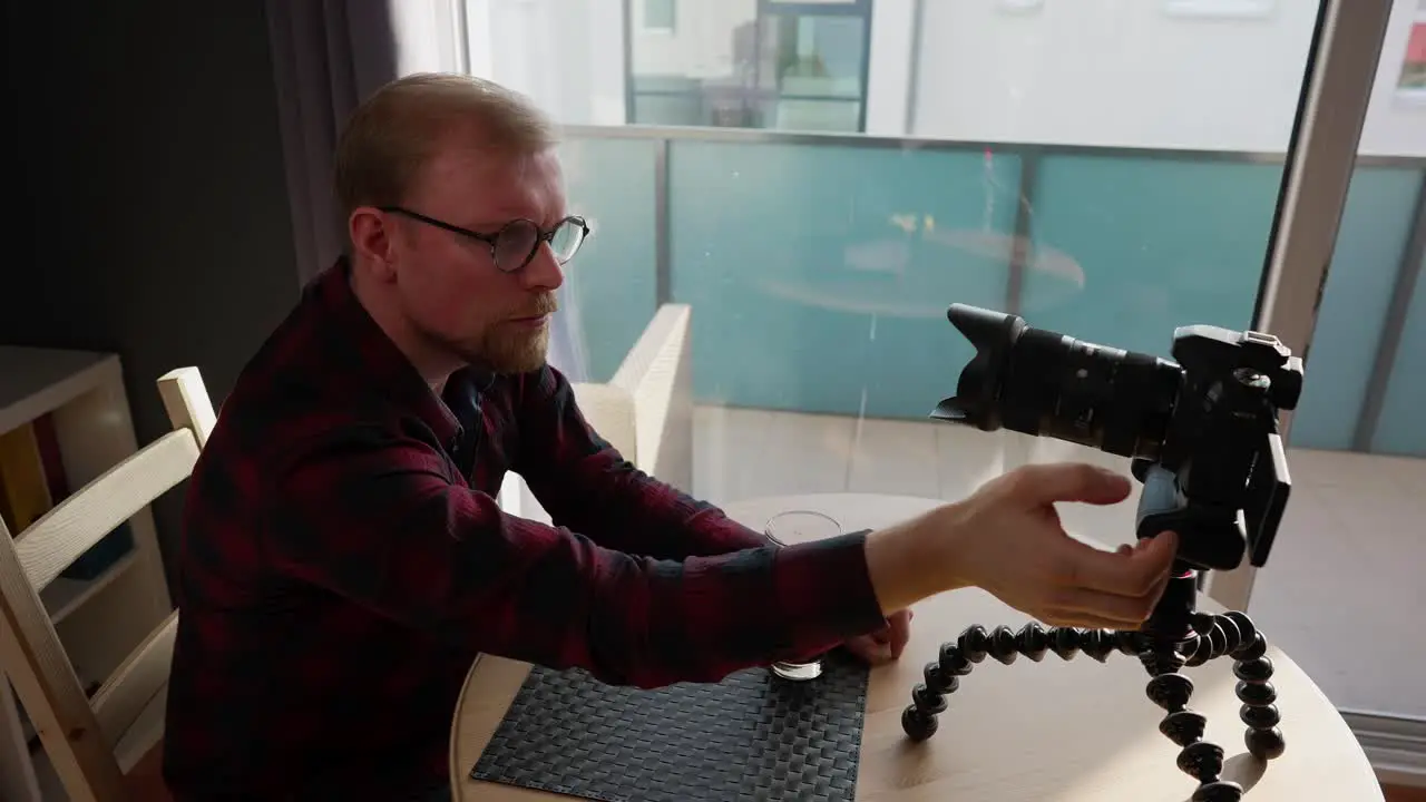 Young Male Vlogger sits at Table Adjusts Camera for Filming Medium Shot