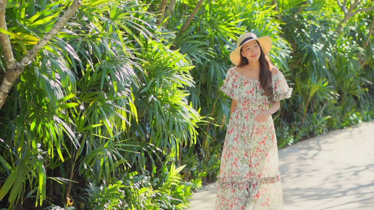 Slow-motion of Asian lady walking towards the camera wearing a long sundress and straw hat on tropical background in Bali