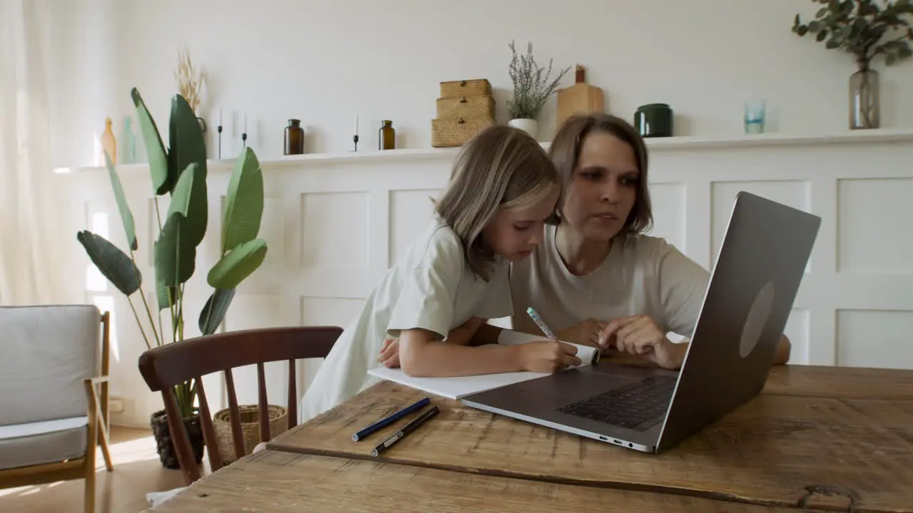 A Pretty Blonde Little Girl Does Her Homework With The Help Of Her Mother While Searching For Information Online On The Laptop