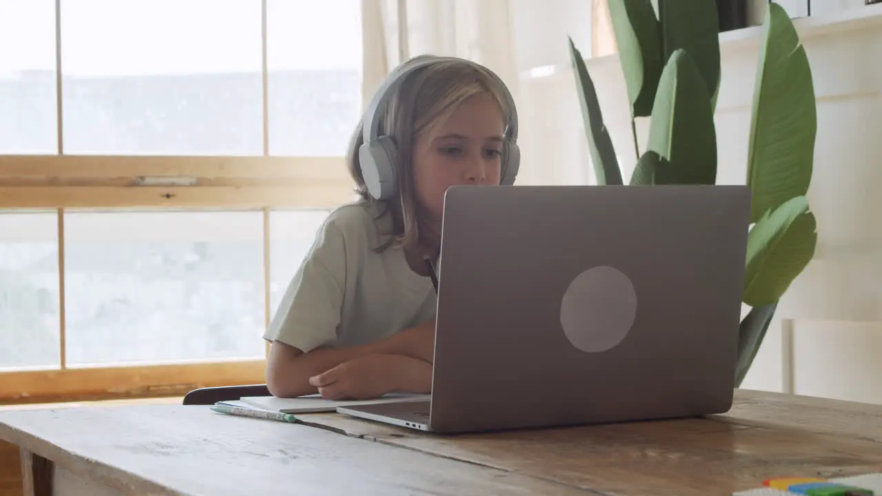 A Pretty And Intelligent Blonde Girl With Headphones Does Her Homework Writing On Paper While Looking At A Laptop Screen