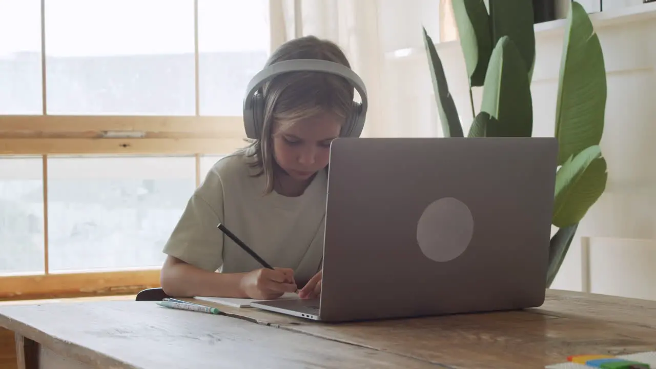 A Pretty And Intelligent Blonde Girl With Headphones Looks At The Laptop Screen And Does Her Homework Using A Pencil And Paper