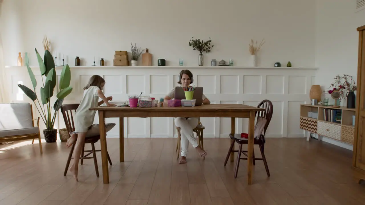 Wide Shot Of A Mother Working At Home Making A Videocall When Her Daughter Comes In And Sits Down Next To Her To Do Her Homework