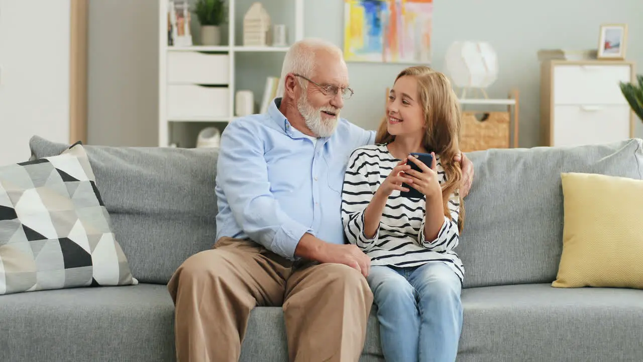 Grandfather Embracing His Pretty Granddaughter While They Sitting On The Sofa In The Living Room And Girl Tapping On The Smartphone Screen And Man Watching At It