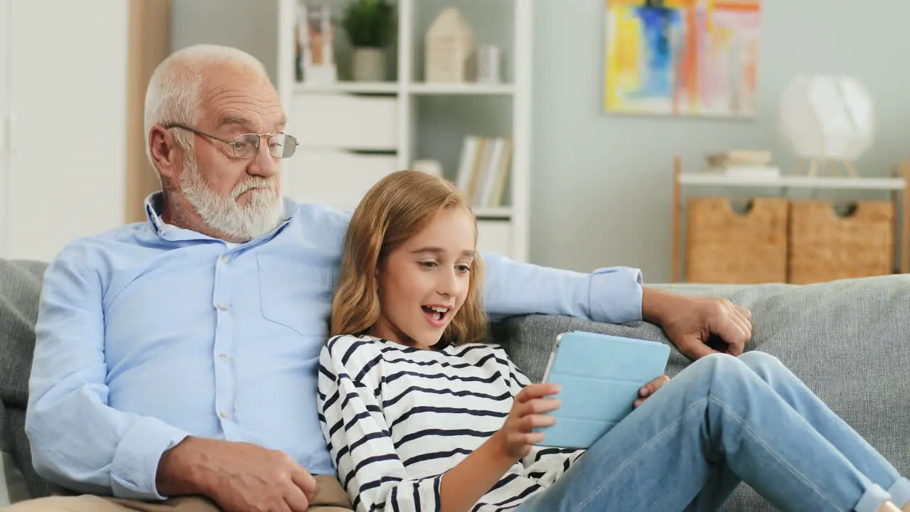 Portrait Shot Of The Excited Grandfather And Granddaughter Watching Video On The Tablet Device While Sitting Together On The Sofa In The Cozy Living Room