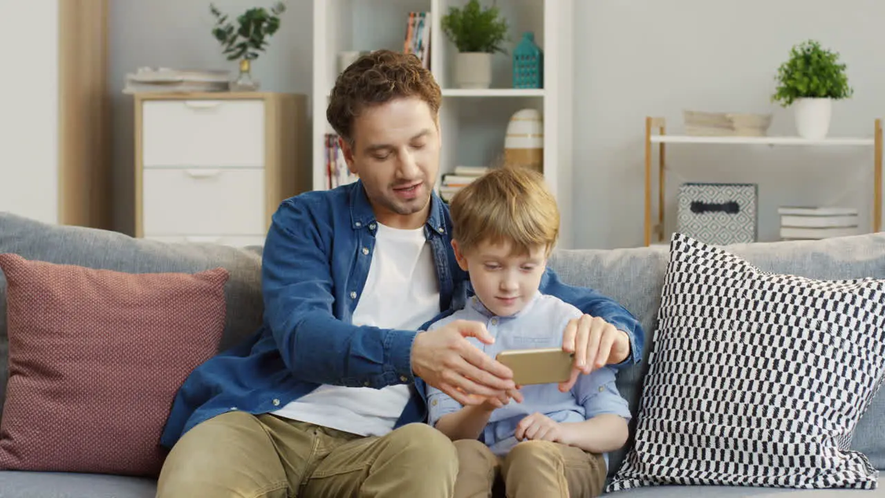 Father And Small Son Spending Time Together Man Teaching A Boy To Use The Smartphone Device At Home In The Living Room