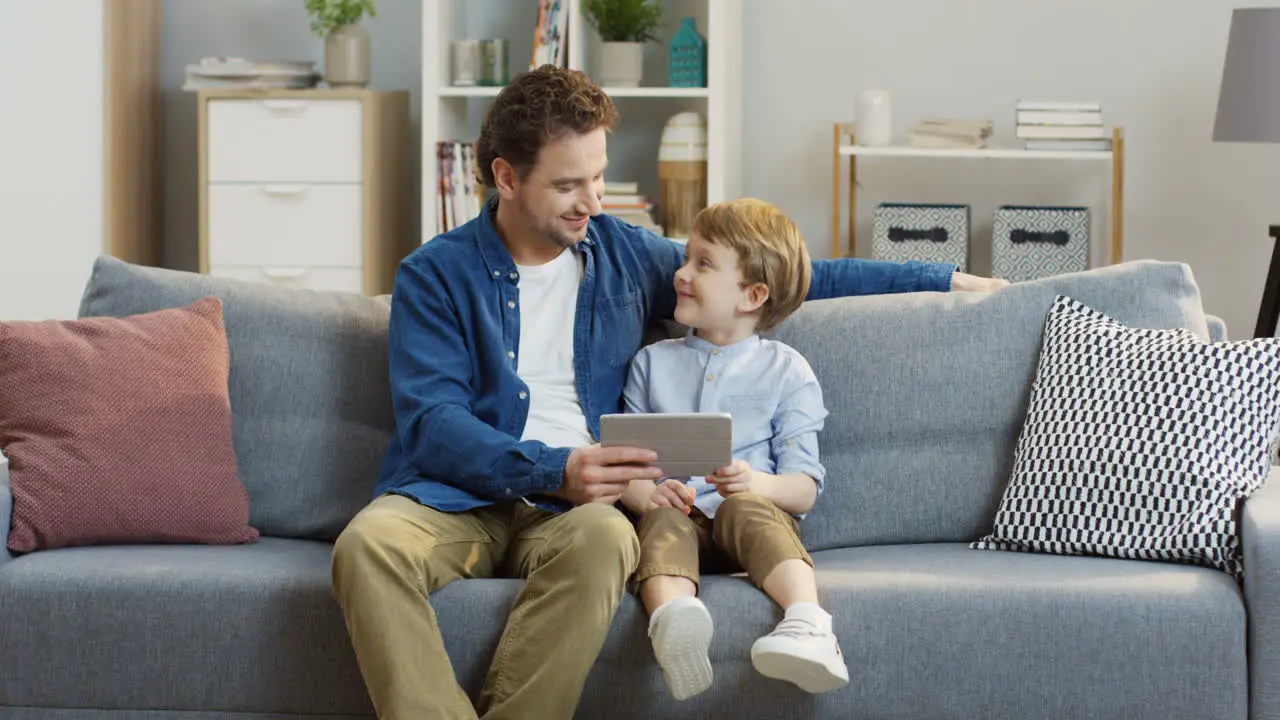 Handsome Father Sitting On The Gray Couch And Hugging His Cute Little Son While They Watching Something On The Tablet Computer In The Living Room