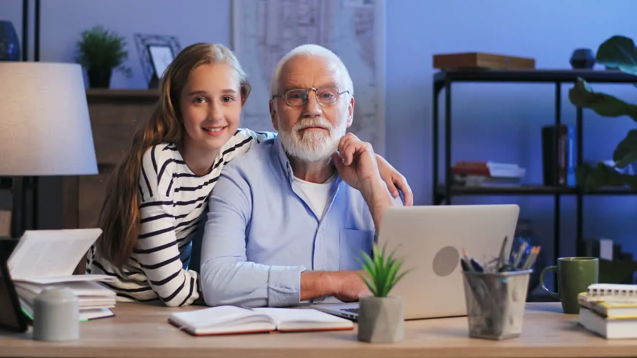 Portrait Of The Pretty Granddaughter Standing At Her Grandpa Who Working Late In The Evening On The Computer Then They Smiling To The Camera