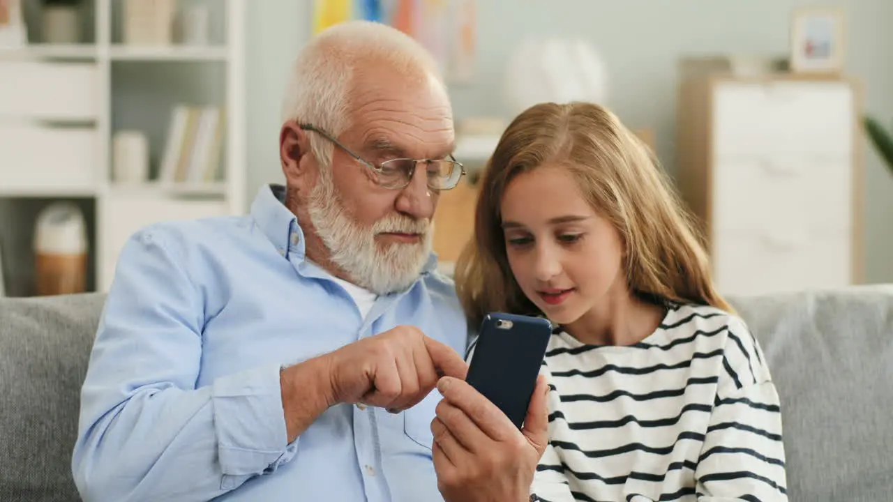 Portrait Of The Cute Blond Teen Girl And His Grey Haired Grandpa Who Showing Her Something On The Smartphone Screen Sitting On The Couch At Home