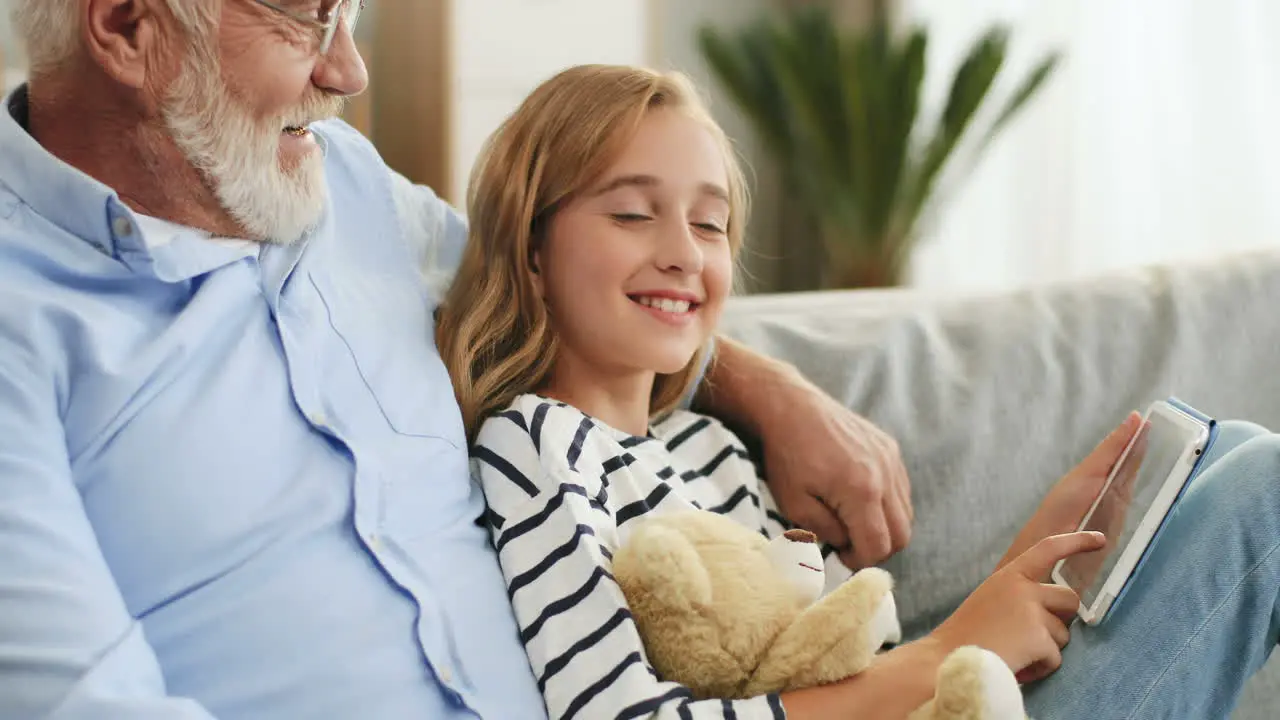 Close Up Of The Senior Grandfather In Glasses Sitting On The Couch And Hugging His Pretty Teen Granddaughter Who Holding Teddy Bear And Scrolling On The Tablet Device