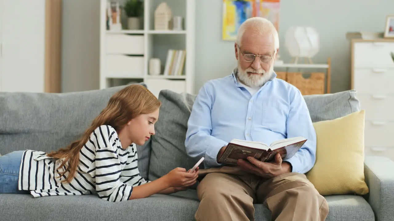 Grey Haired Old Grandfather In The Glasses Reading A Book And His Granddaughter Using Smartphone While They Resting On The Couch