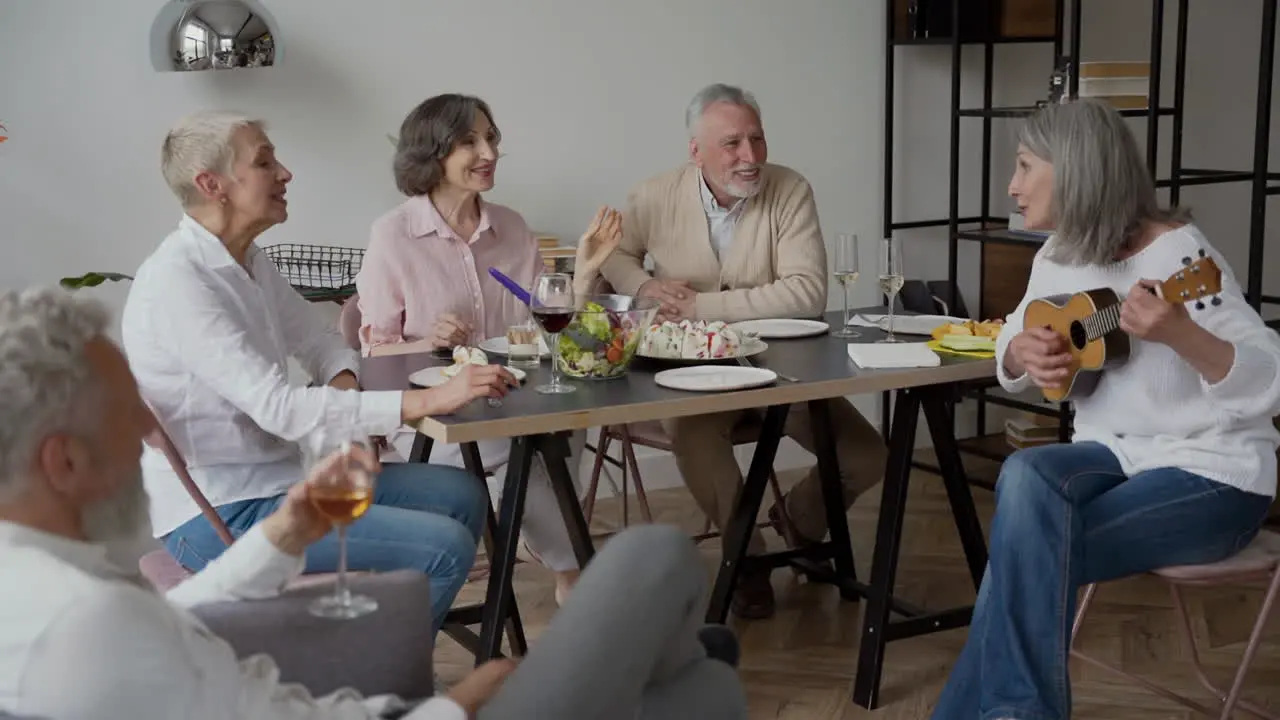 Happy Senior Woman Singing And Playing The Guitar Sitting On Chair While Her Elderly Friends Listening To Her And Singing Together Sitting At The Table