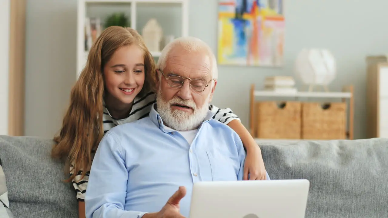 Pretty Teen Girl Hugging Her Grandfather From Behind While He Sitting On The Sofa And Typing On The Laptop At Home