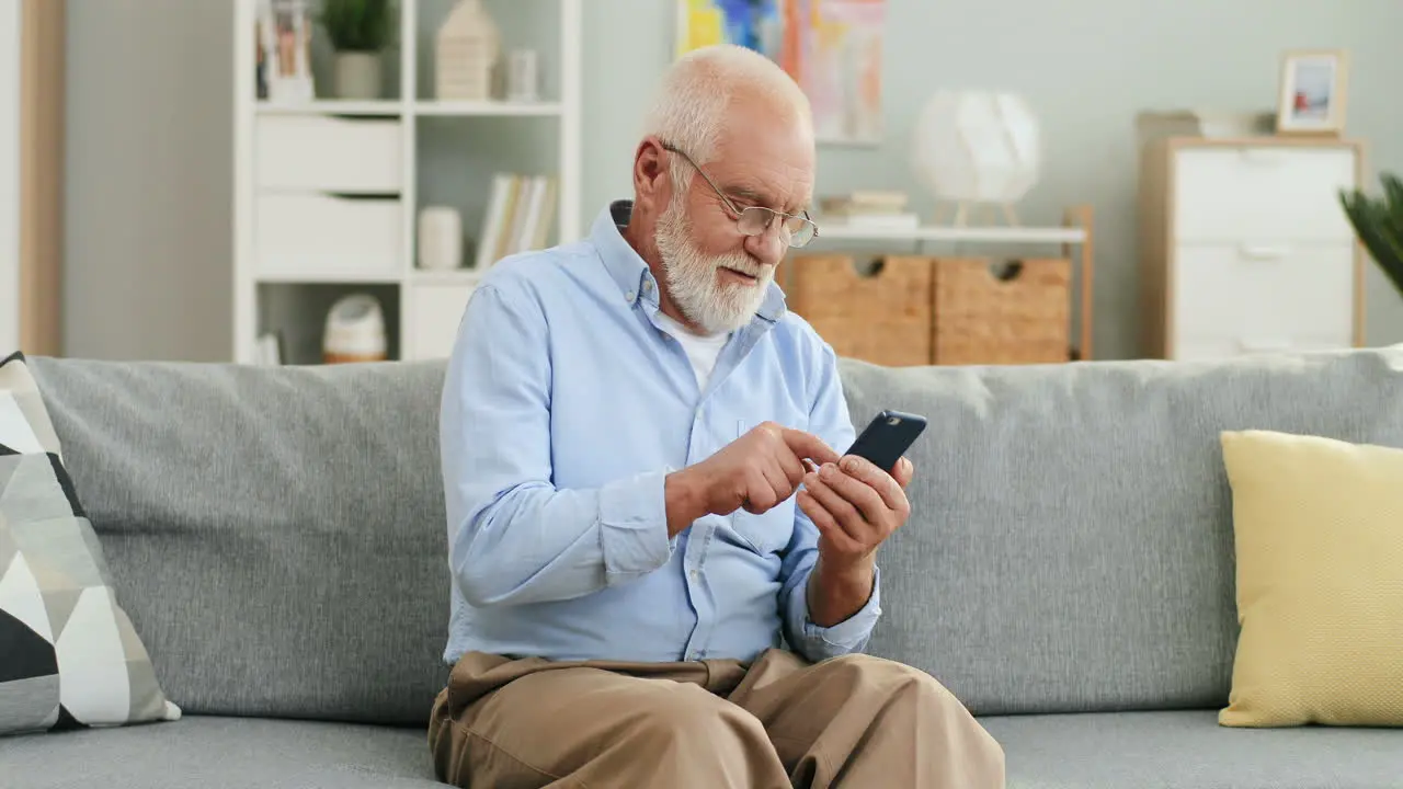 Old Grey Haired Man In Glasses Sitting On The Couch At Home And Texting A Message On The Smartphone