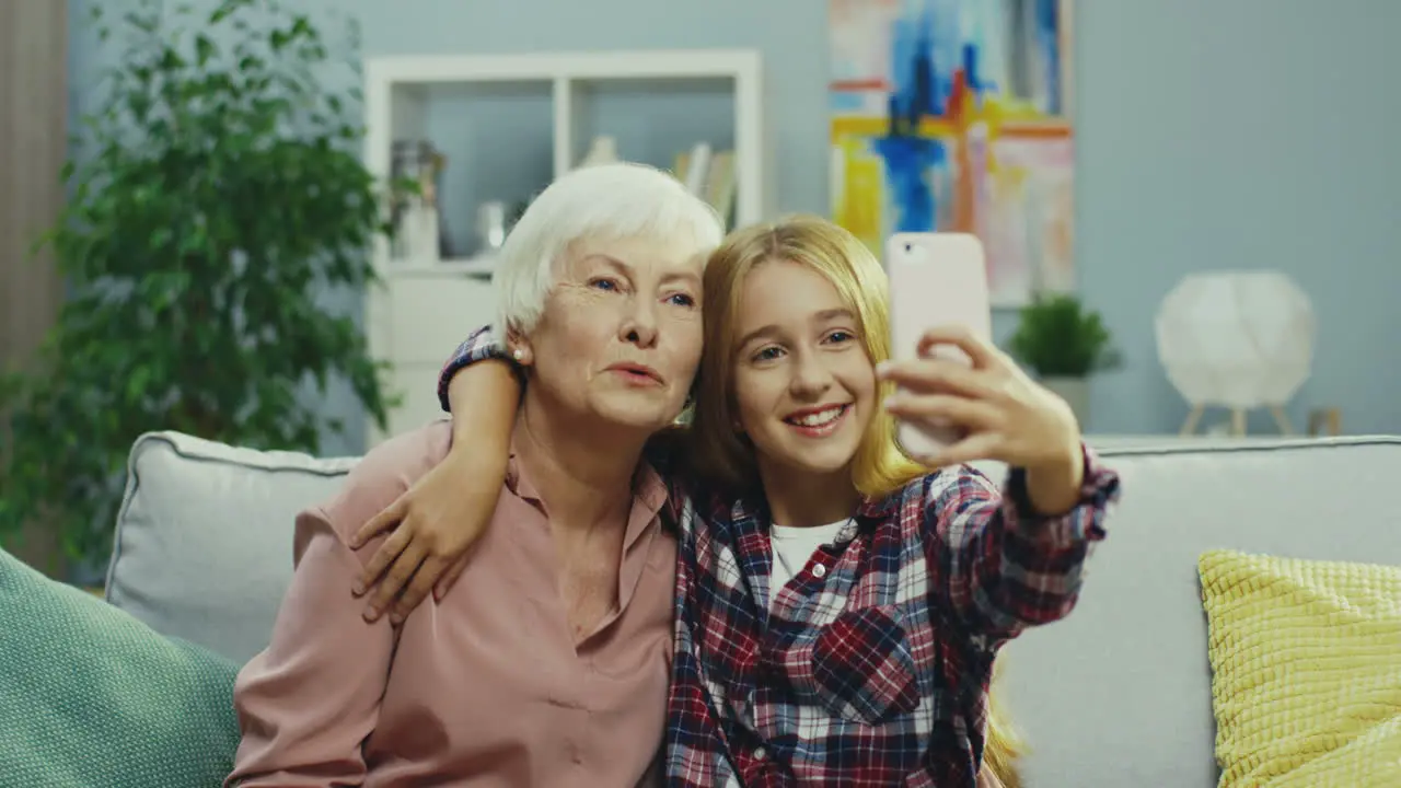 Close Up Of The Cute Teen Girl And Her Smiled Gray Haired Grandmother Posing To The Smartphone Camera While Taking Selfie Photo On The Couch At Home