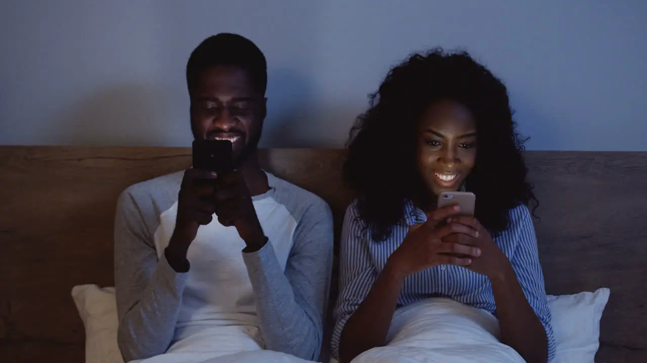 American Couple Laughing While Looking At Their Cell Phones In Bed Before Bedtime