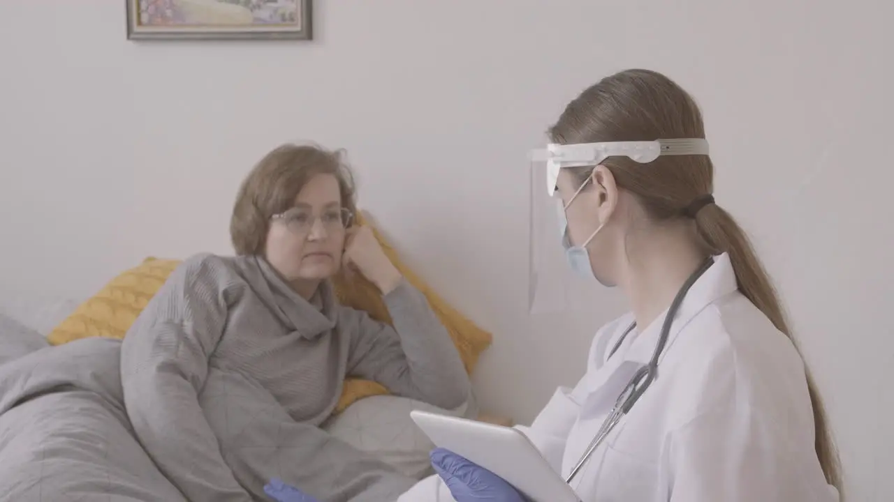 Senior Woman Sitting On Bed While Talking To Female Doctor In Medical Mask And Protective Screen Using A Table