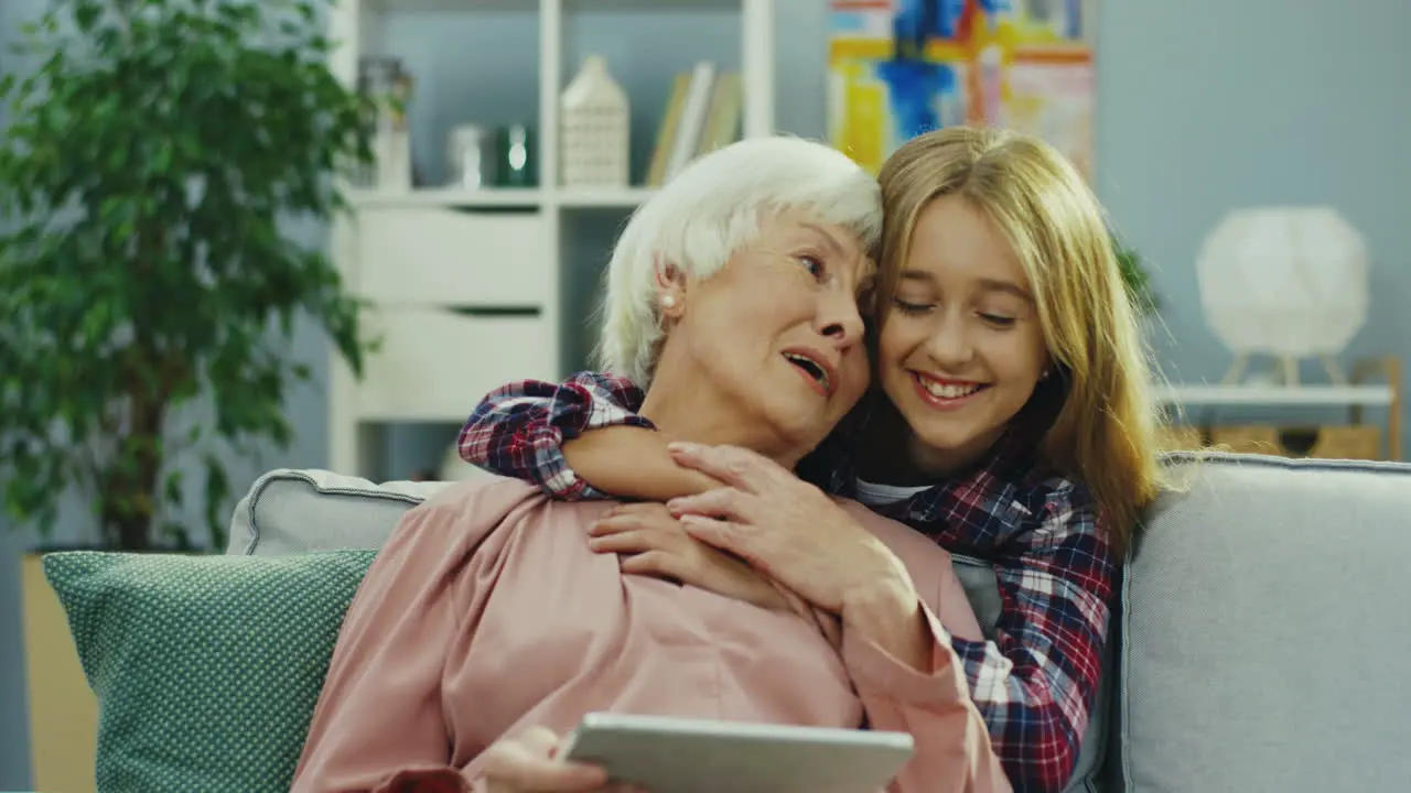 Gray Haired Senior Woman Sitting On The Couch With Tablet Device And Her Cute Teenage Granddaughter Coming From Behind And Hugging Her