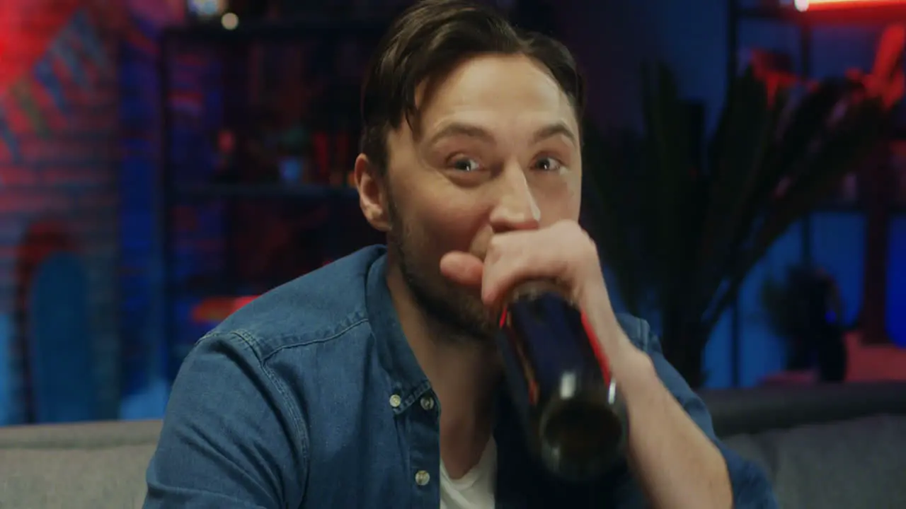 Portrait Shot Of The Handsome Young Guy Smiling Cheerfully To The Camera And Drinking Beer In The Dark Room