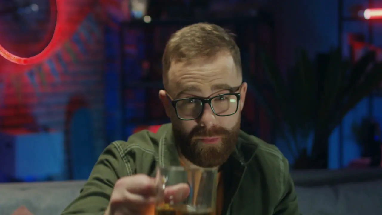 Portrait Of The Young Cheerful Man In Glasses Drinking Beer And Doing Cheers Gesture To The Camera And Smiling In The Dark Room