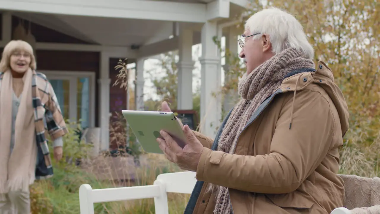 An Older Couple Outside Their Home The Man Uses A Table While Making A Video Call And Invites His Wife To Join