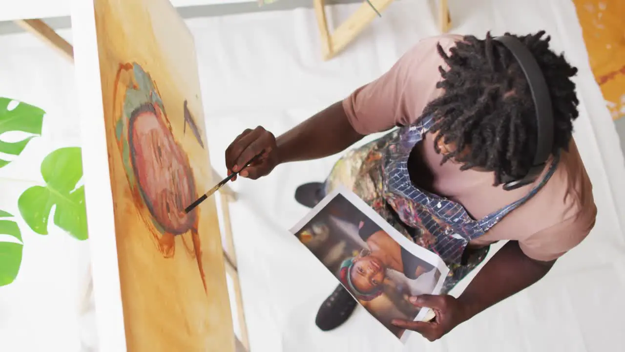 Overhead view of african american male artist painting on canvas looking at photo at art studio