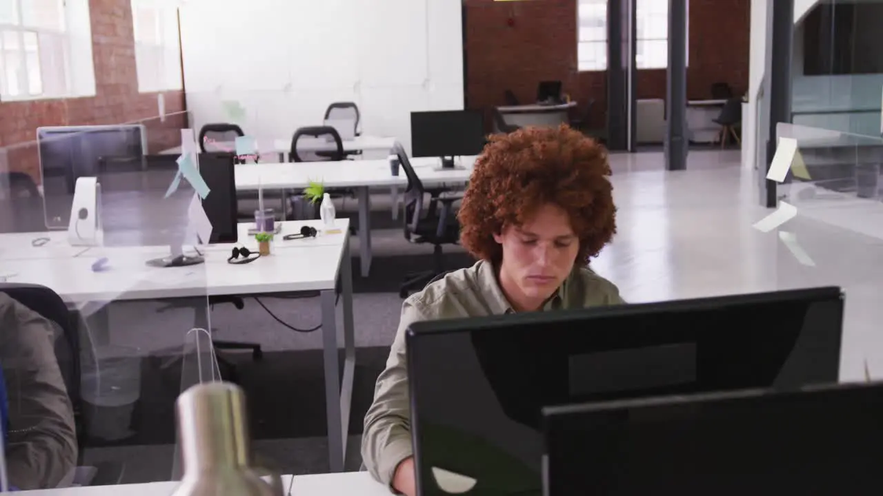 Mixed race businessman sitting at desk with sneeze guard using desktop computer looking at screen