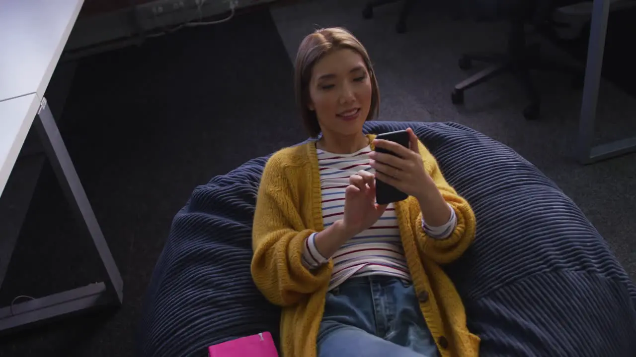 Happy asian business woman lying on bean bag using smartphone during break