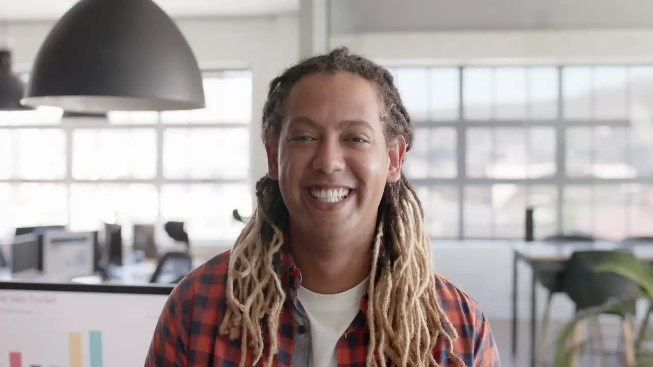 Portrait of happy biracial businessman with dreadlocks smiling in creative office in slow motion