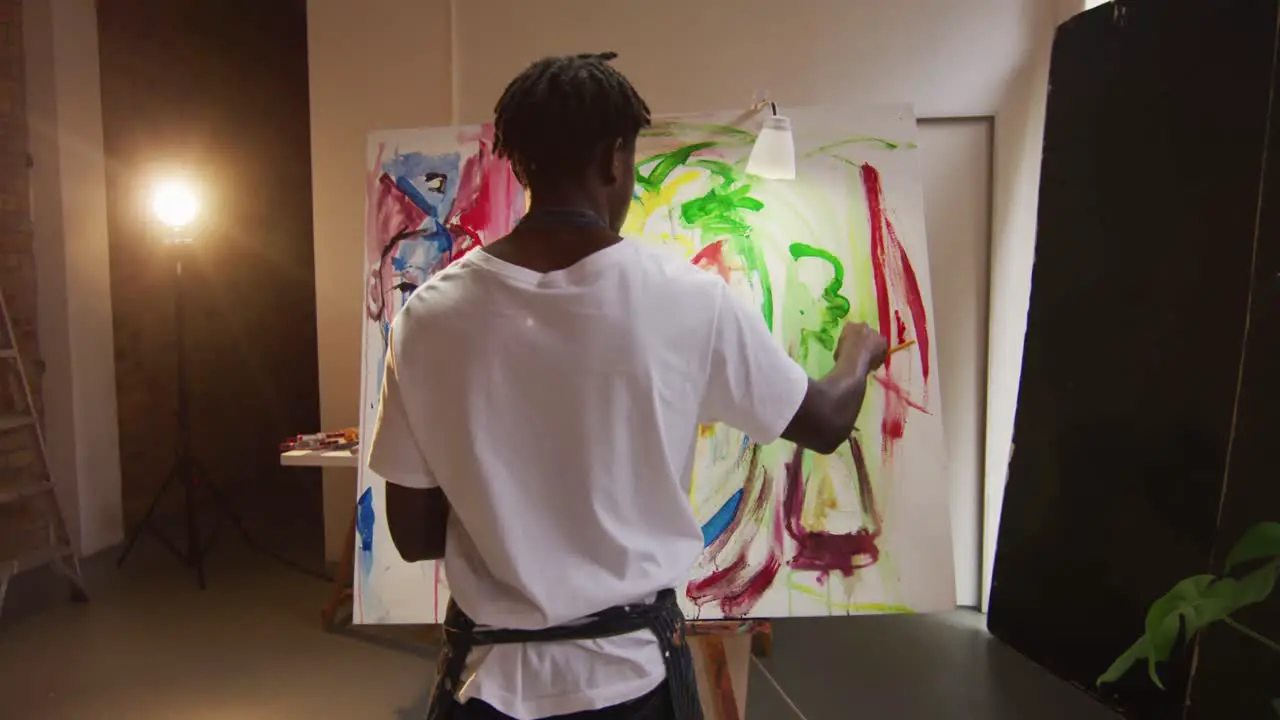 African american male artist wearing apron painting with paint brush on canvas at art studio