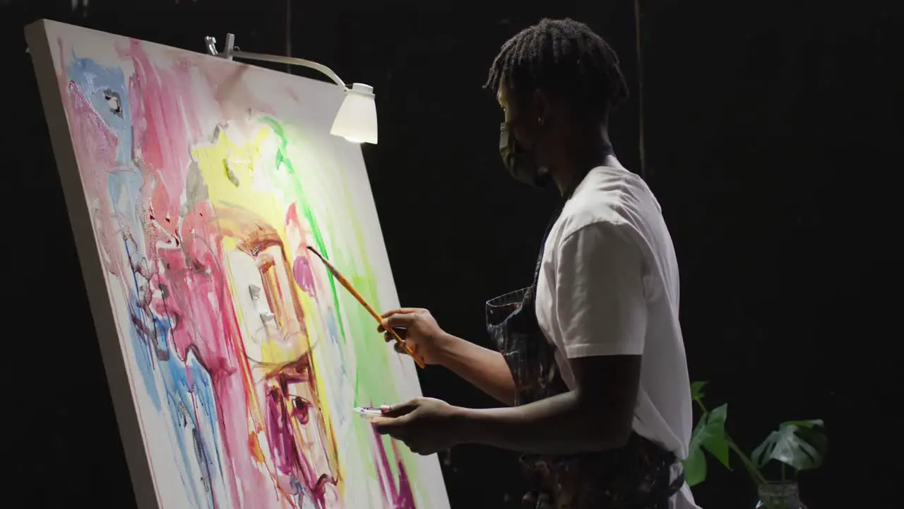 African american male artist wearing face mask painting on canvas at art studio