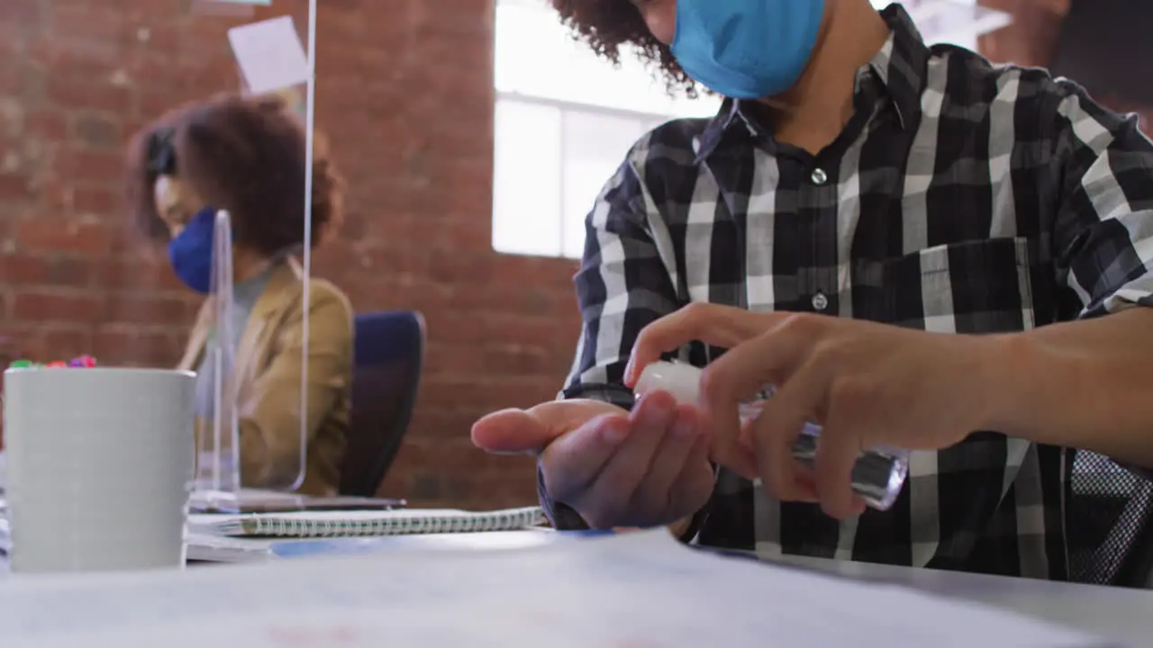 Mixed race businessman disinfecting hands sitting in front of computer wearing face mask