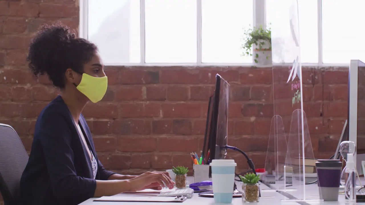 Mixed race businesswoman typing sitting in front of computer wearing face mask