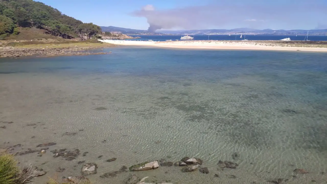 Fire on the horizon behind the coast with forest and beach with luxury yachts anchored on a sunny summer day rolling crane shot upwards Cíes Islands Pontevedra Galicia Spain