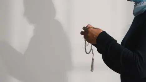 Muslim Woman Wearing Hijab Praying Holding Prayer Beads Casting Shadow On Wall Behind