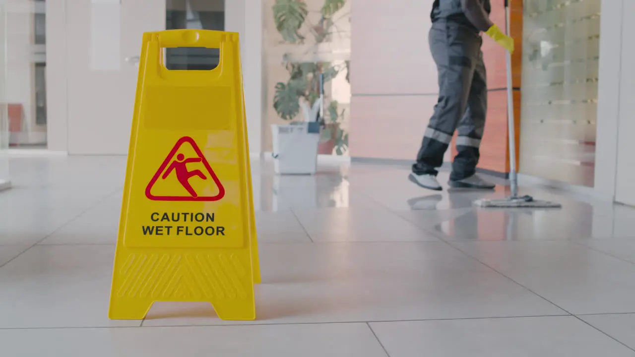 Cleaning Man Cleaning The Floor With Mop Inside An Office Building Behind A Wet Floor Warning Sign