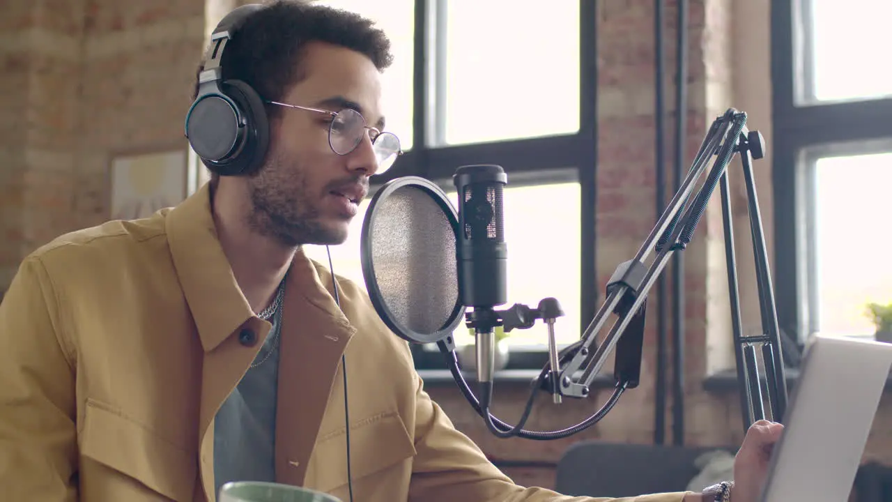 Man Recording A Podcast Wearing Eyeglasses And Headphones Talking Into A Microphone While Sitting On A Table With Laptop