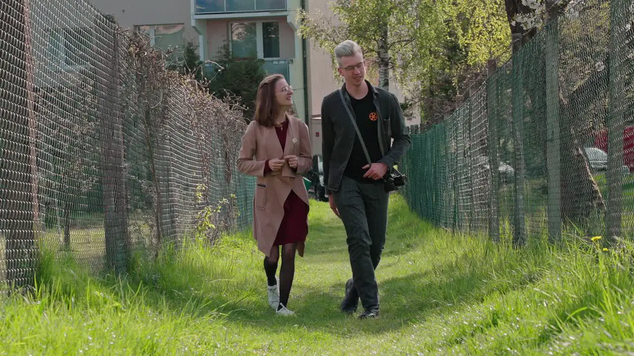 Couple of young man and girl walking between fences on green spring grass in sunny spring